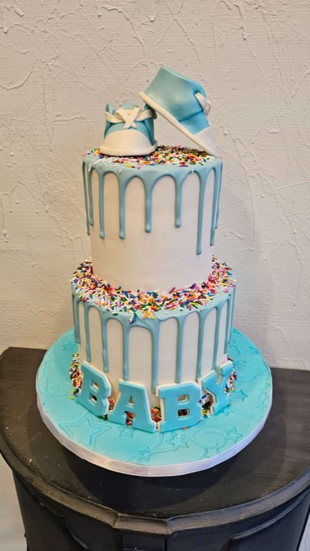 White and baby blue fondant gender reveal baby shower cake with blue drip, BABY in blue letters, colorful sprinkles, and blue baby bib and booties