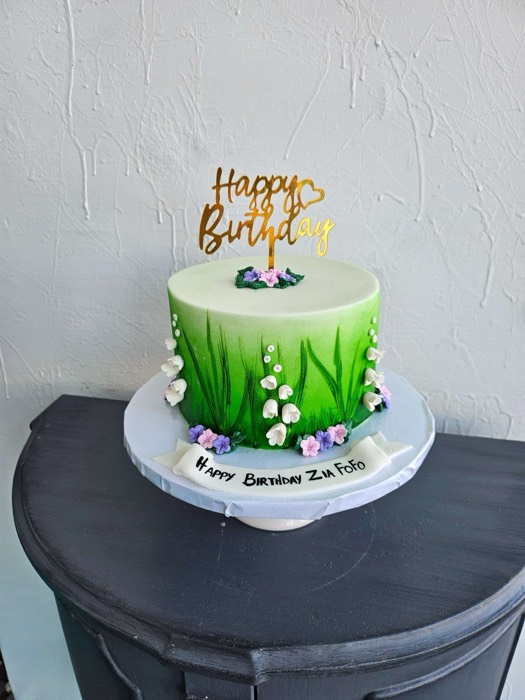 Green ombre painted birthday cake with white lily of the valley bells, purple and pink blossoms, pearl balls, gold Happy Birthday topper, and Happy Birthday banner