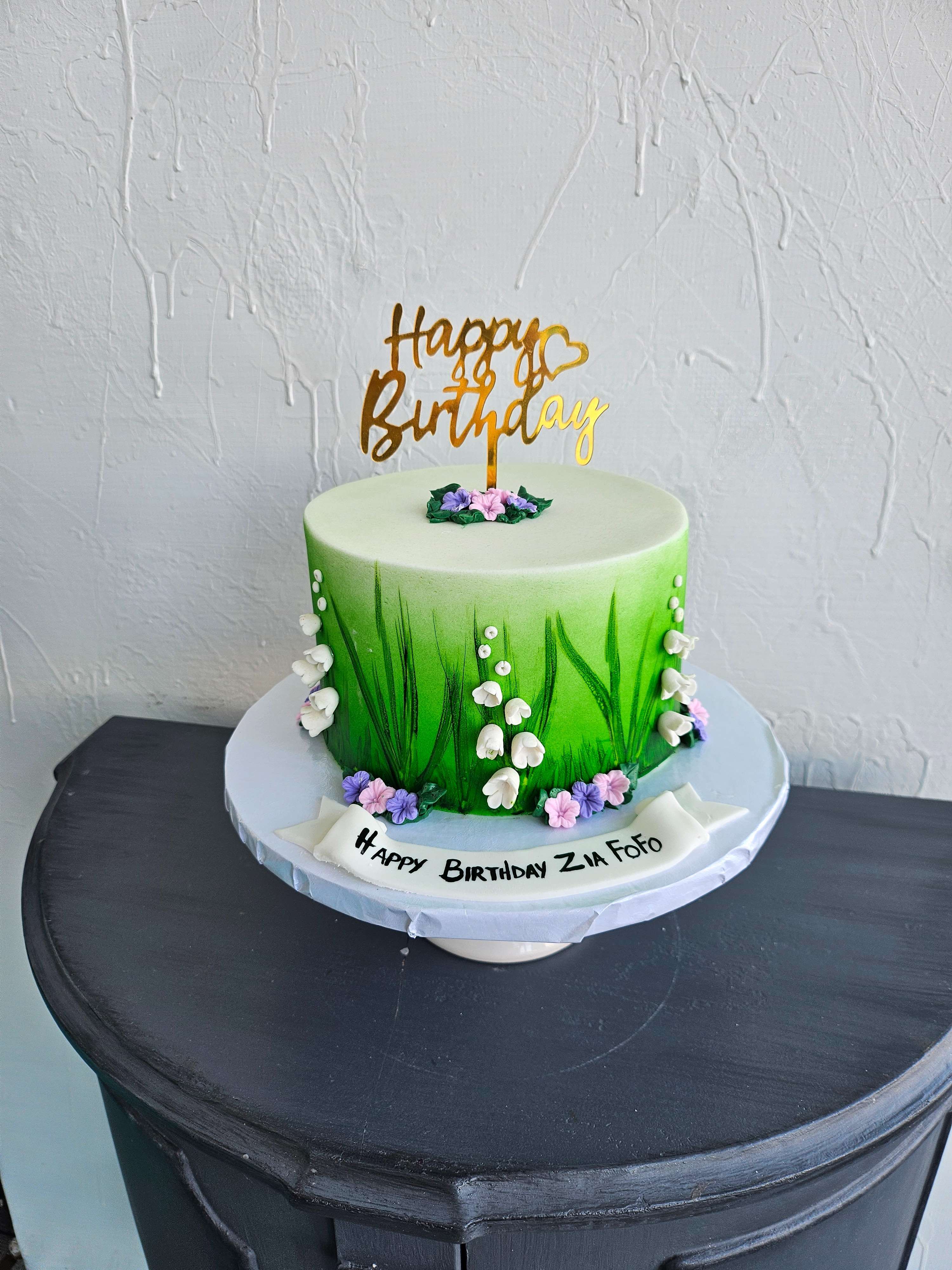 Green ombre painted birthday cake with white lily of the valley bells, purple and pink blossoms, pearl balls, gold Happy Birthday topper, and Happy Birthday banner