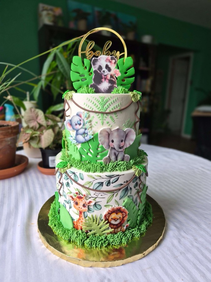 Two-tier jungle safari baby shower cake with edible printed giraffe, lion, elephant, panda, and mouse characters on white fondant, green tropical leaf accents, green grass borders, and gold Baby hoop topper