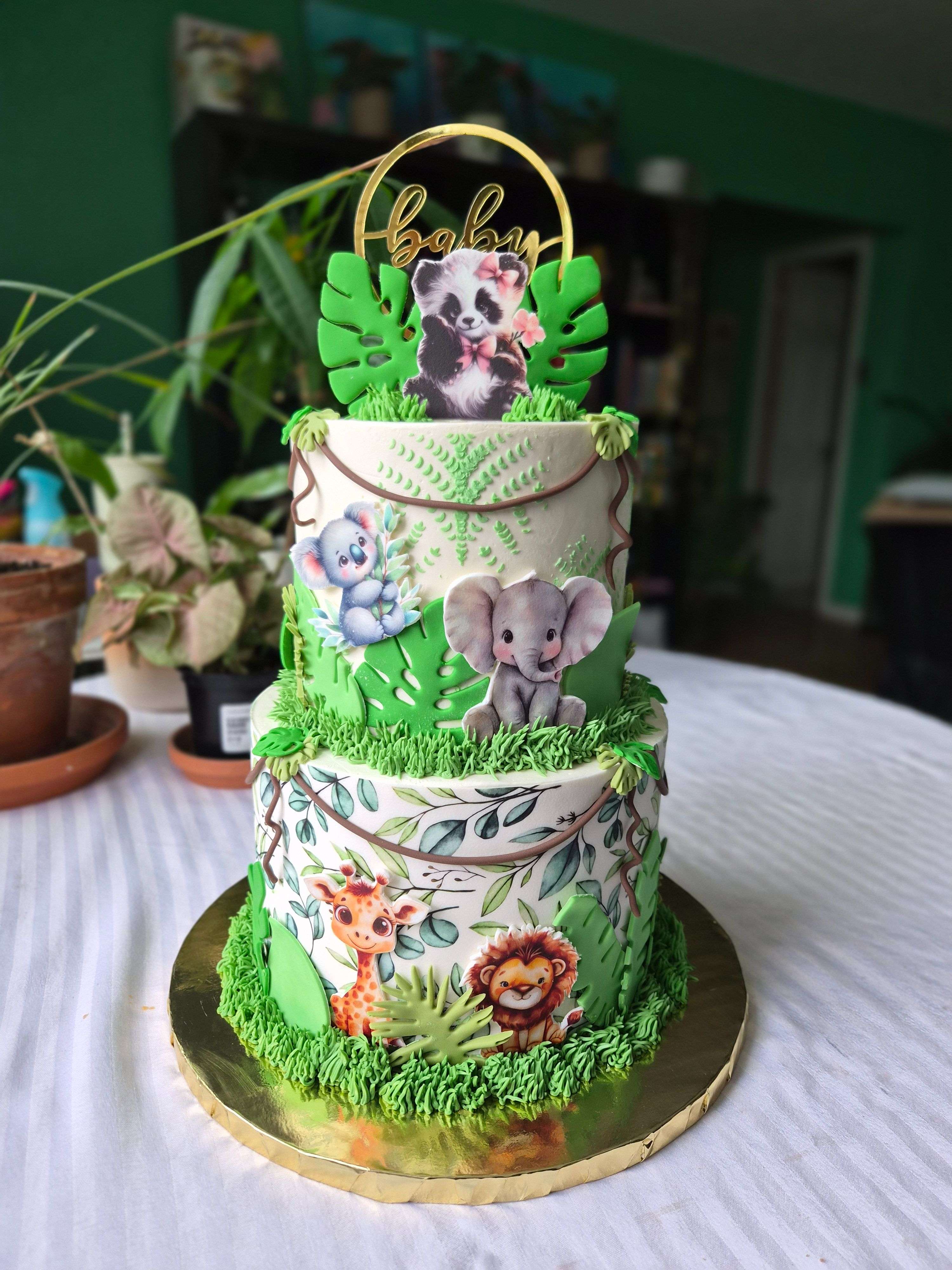 Two-tier jungle safari baby shower cake with edible printed giraffe, lion, elephant, panda, and mouse characters on white fondant, green tropical leaf accents, green grass borders, and gold Baby hoop topper