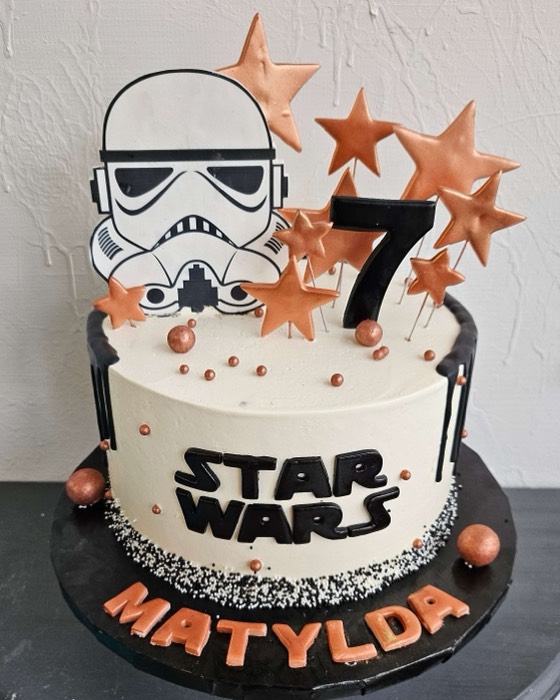 White buttercream Star Wars 7th birthday cake with black Star Wars logo, black drip, Stormtrooper helmet topper, black number 7, copper star sprinkles, and orange personalized name letters