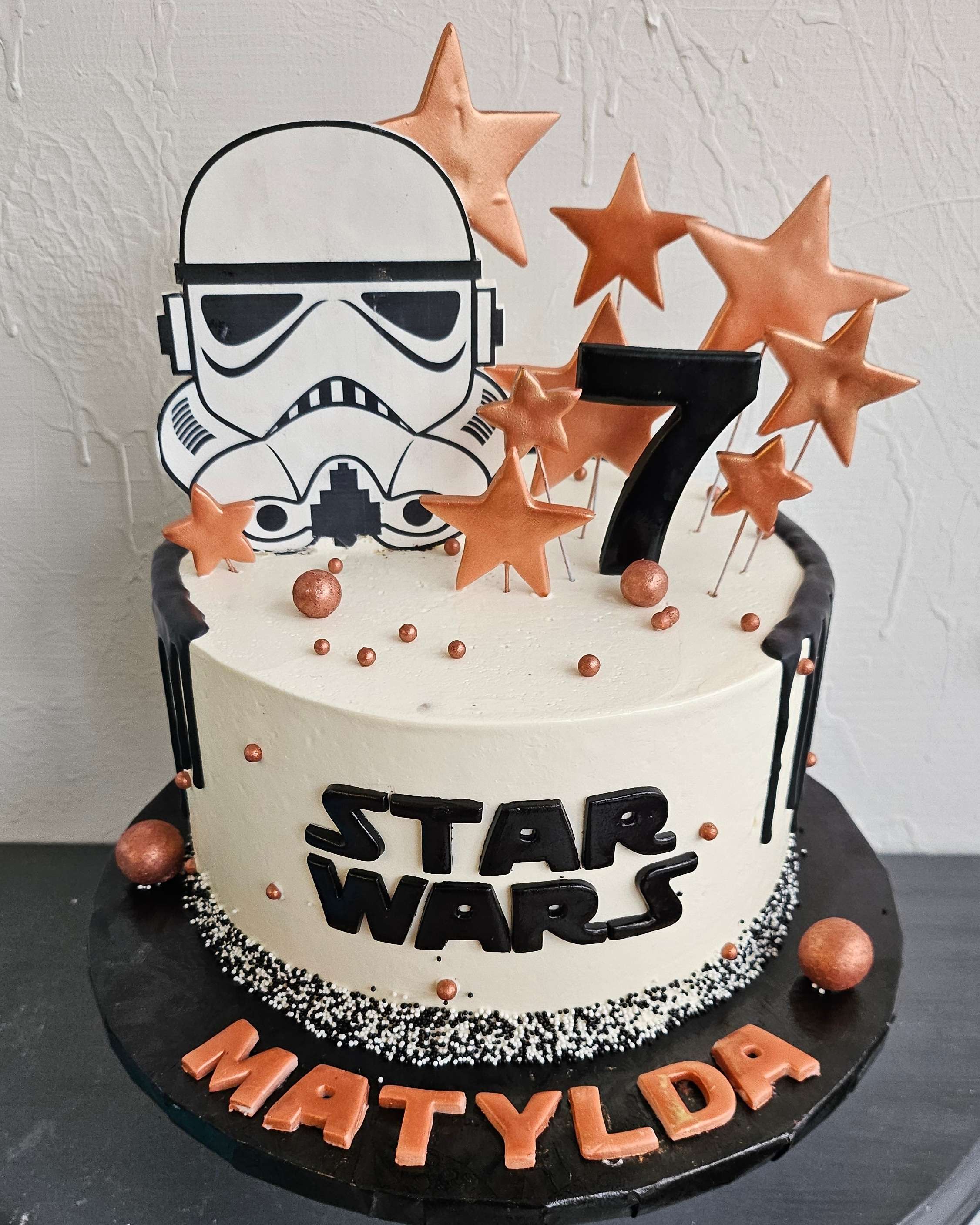 White buttercream Star Wars 7th birthday cake with black Star Wars logo, black drip, Stormtrooper helmet topper, black number 7, copper star sprinkles, and orange personalized name letters