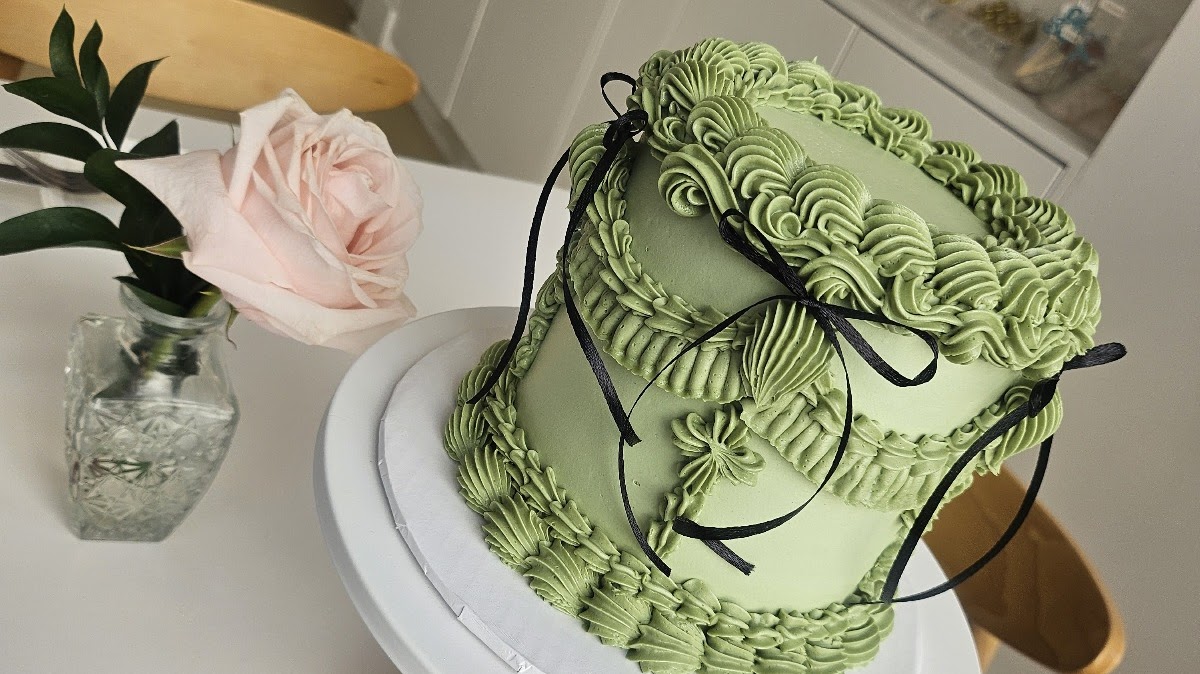 Sage green two-tier lambeth-style cake with elaborate piped ruffles, fan motifs, and black satin ribbon bows, on a white cake stand